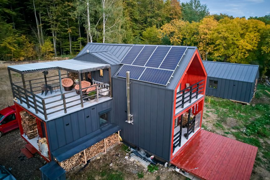 Modern off-grid home with rooftop solar panels, outdoor deck seating, and a forest backdrop